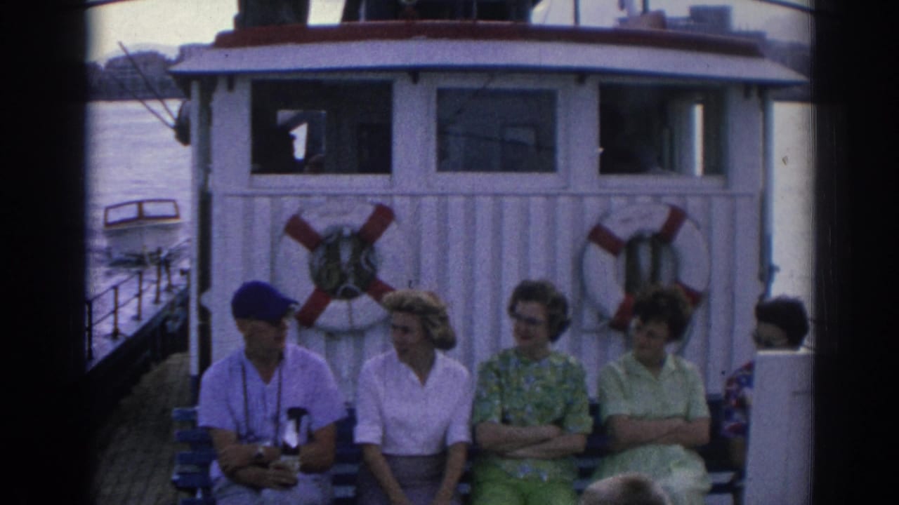 Adults in 1960s clothing sit together on an Izmir ferry deck, including a man in a blue cap, with water and shoreline visible.