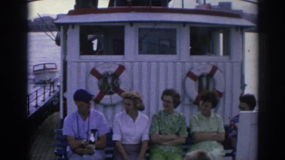Adults in 1960s clothing sit together on an Izmir ferry deck, including a man in a blue cap, with water and shoreline visible.