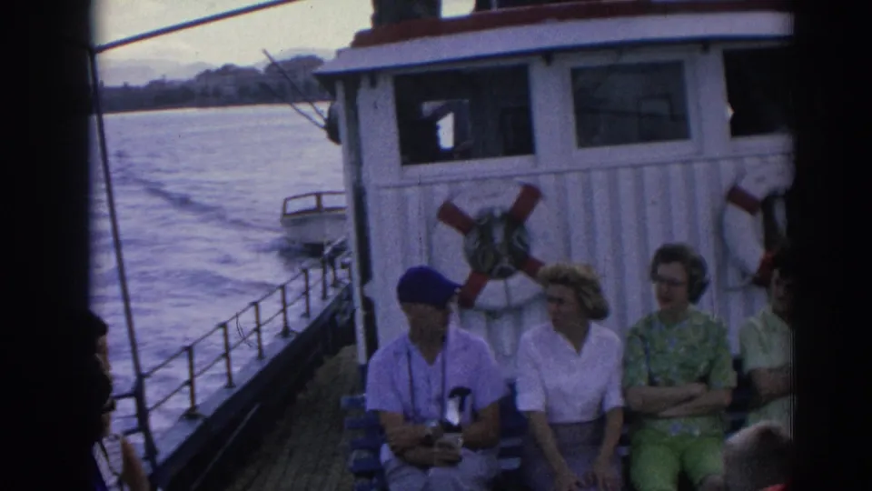 Izmir ferry passengers sit near the cabin and railing while the moving boat leaves a wake across the water.