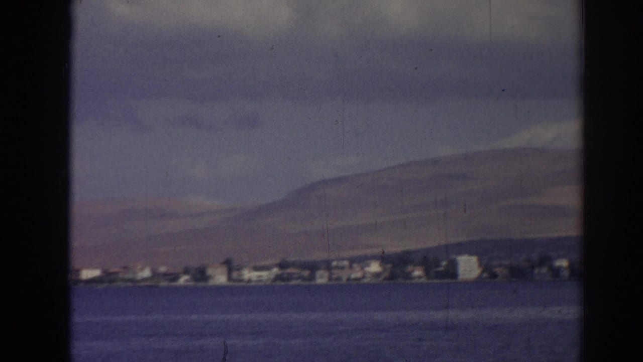 1962 Izmir home-movie frame showing a distant shoreline across blue water, backed by low hills and soft clouds.