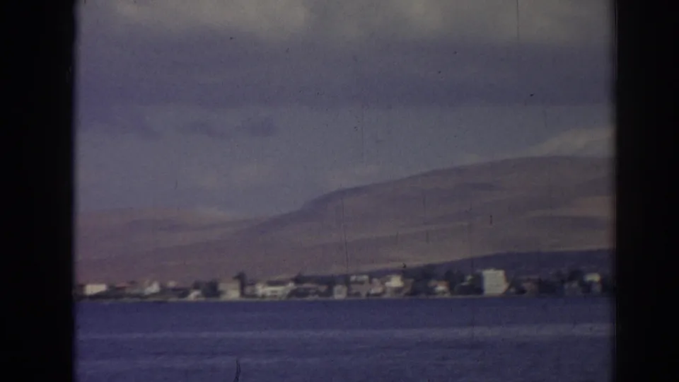 1962 Izmir home-movie frame showing a distant shoreline across blue water, backed by low hills and soft clouds.