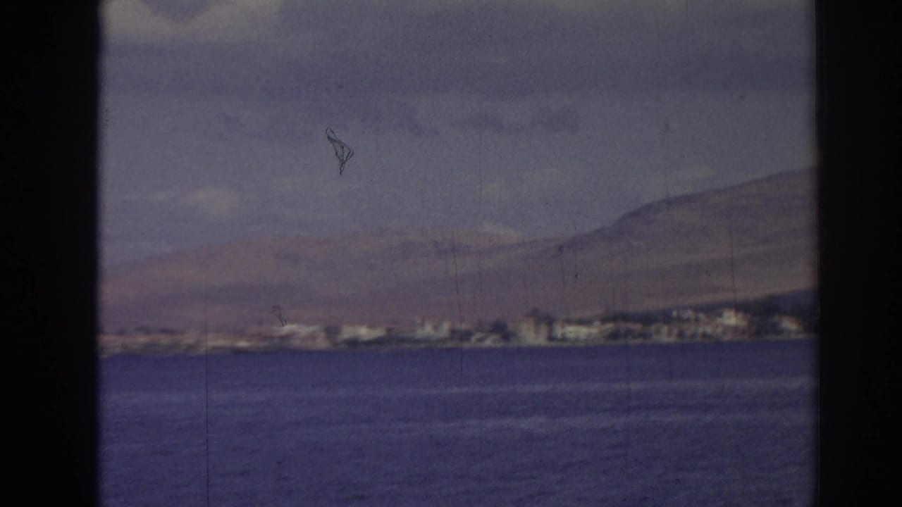 1962 Izmir coastal view with calm water in the foreground, a distant built shoreline, and hills under pale clouds.