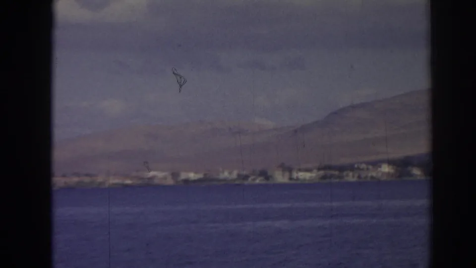 1962 Izmir coastal view with calm water in the foreground, a distant built shoreline, and hills under pale clouds.