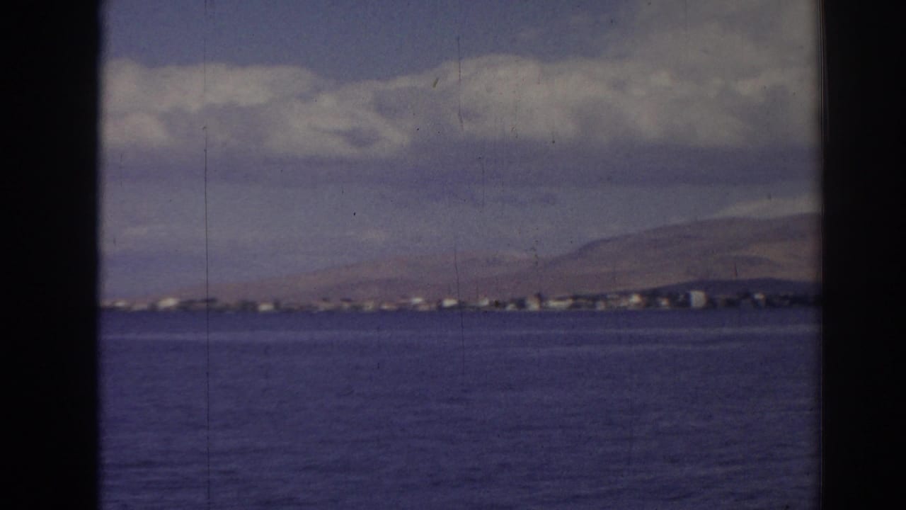 Wide 1962 Izmir shoreline frame with blue water, a thin line of coastal buildings, hills, and puffy clouds.