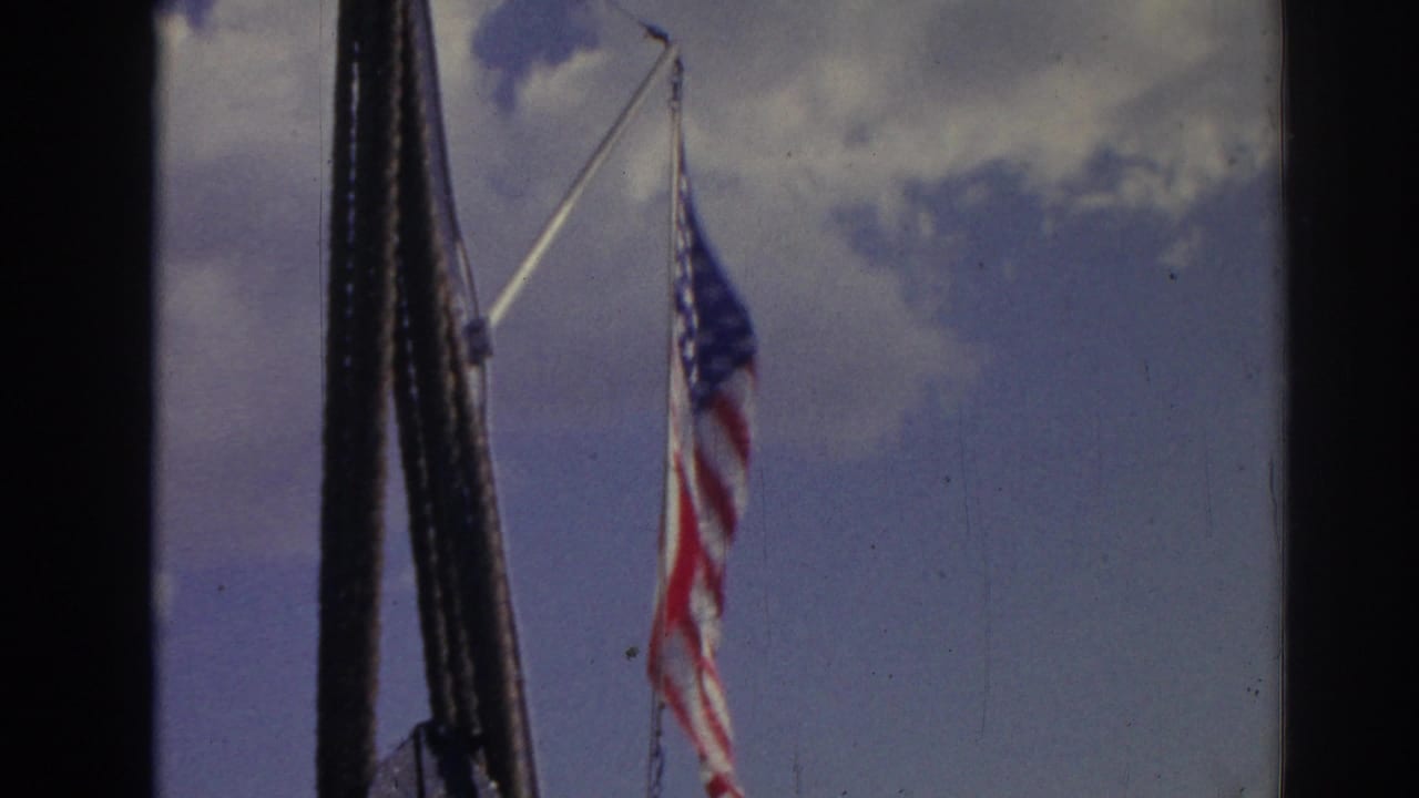 1962 Izmir home-movie frame showing a U.S. flag near a mast or pole, with blue sky and clouds behind it.