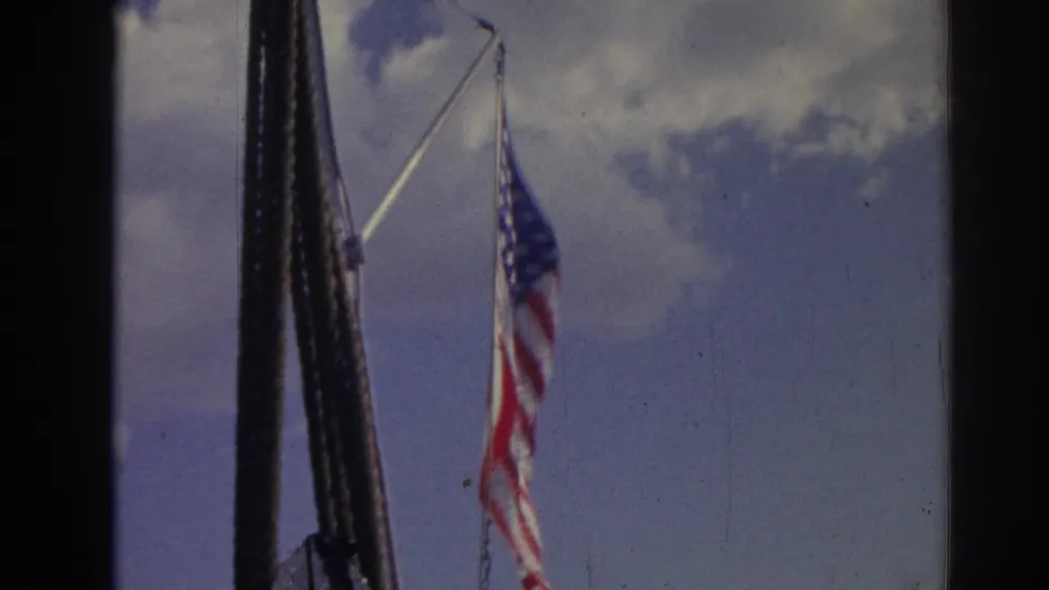 1962 Izmir home-movie frame showing a U.S. flag near a mast or pole, with blue sky and clouds behind it.