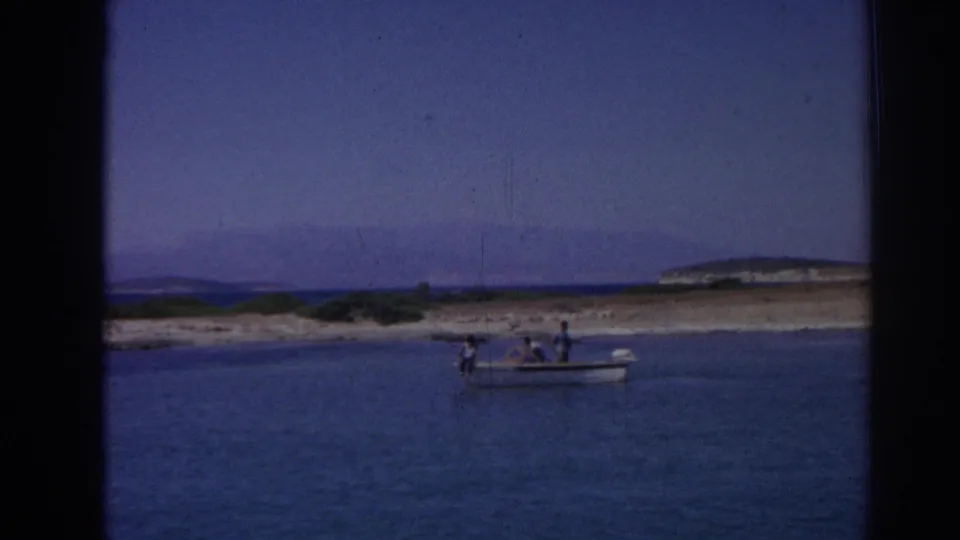 Small motorboat with distant passengers on blue water near Izmir, Turkey, with low coastal land and hills in the background.