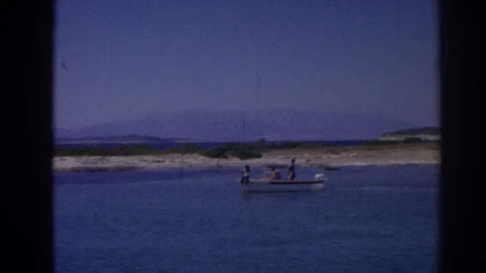 Distant 1962 home-movie view of a small boat on coastal water near Izmir, with passengers visible and hills beyond the shore.