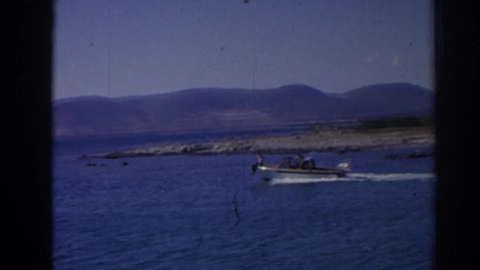1962 Izmir-area coastal frame showing a small motorboat underway on rippled blue water, with passengers and distant hills visible.