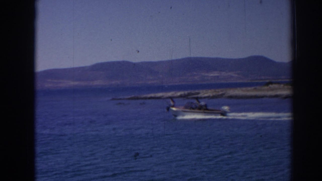 Small white motorboat crossing blue water near a low rocky shoreline.