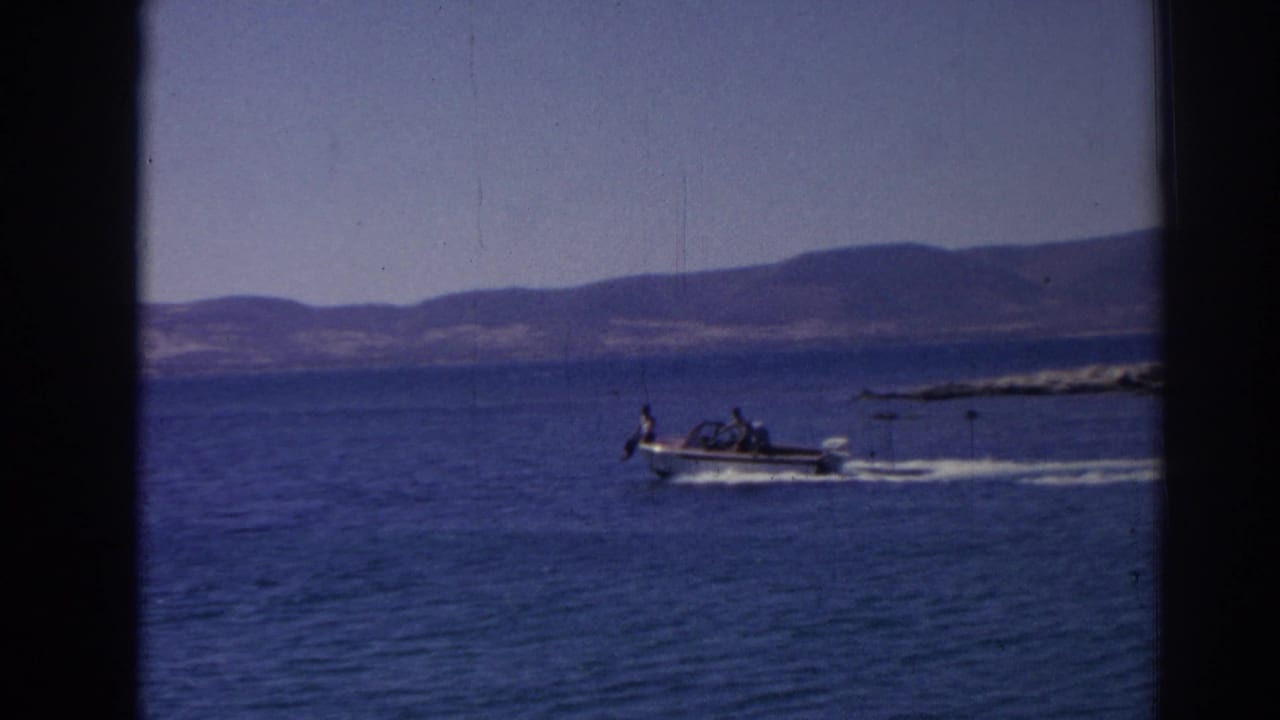 The white motorboat continues along the Izmir-area shoreline, with passengers visible at a distance and a bright wake trailing behind it.