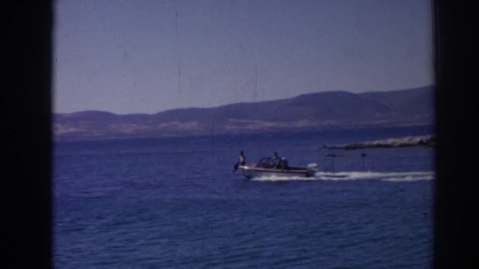 The white motorboat continues along the Izmir-area shoreline, with passengers visible at a distance and a bright wake trailing behind it.