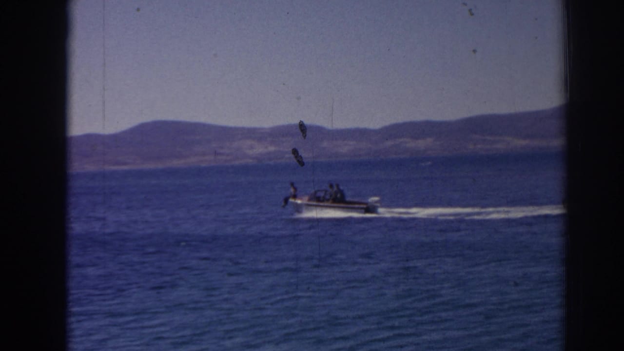A distant small motorboat passes across blue water in a 1962 Izmir coastal home movie, framed by low hills and visible 8mm film marks.