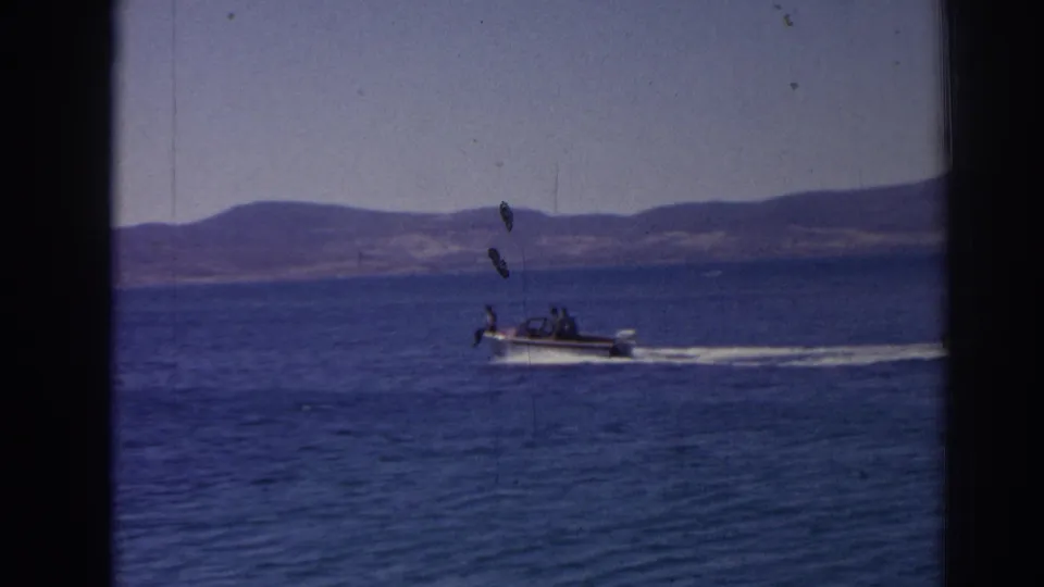 A distant small motorboat passes across blue water in a 1962 Izmir coastal home movie, framed by low hills and visible 8mm film marks.