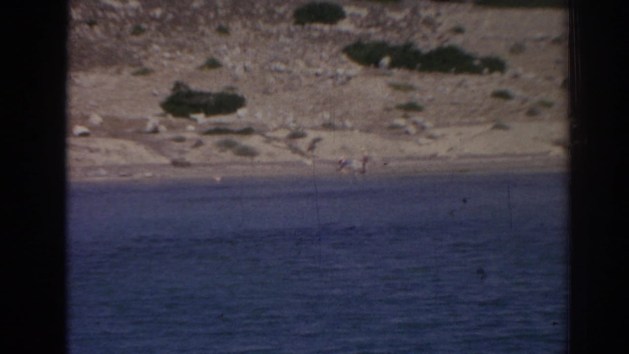 The frame shifts toward the rocky, arid shoreline near Izmir, with blue water in the foreground and small distant figures or objects hard to identify.