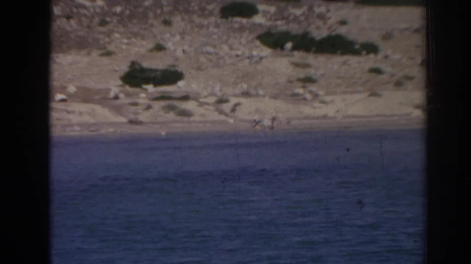 The frame shifts toward the rocky, arid shoreline near Izmir, with blue water in the foreground and small distant figures or objects hard to identify.
