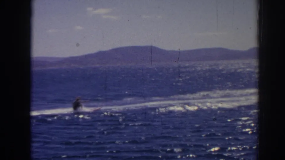 A distant water skier crosses sparkling blue water in 1962 Izmir footage, with hills visible along the far shore.