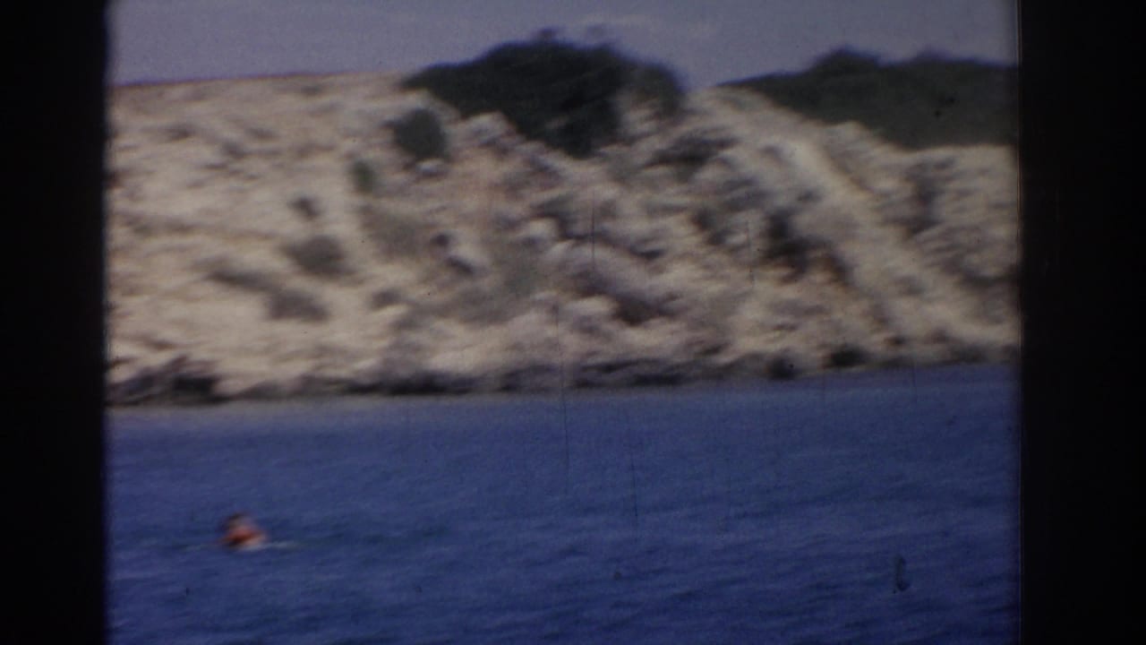 1962 Izmir home-movie frame showing a distant swimmer's head in blue water below a soft, rocky coastal slope.