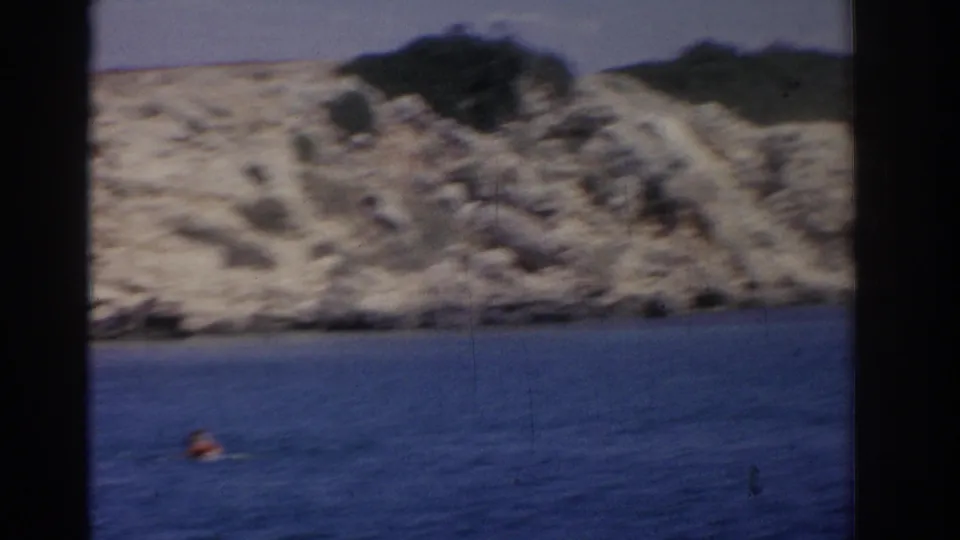1962 Izmir home-movie frame showing a distant swimmer's head in blue water below a soft, rocky coastal slope.