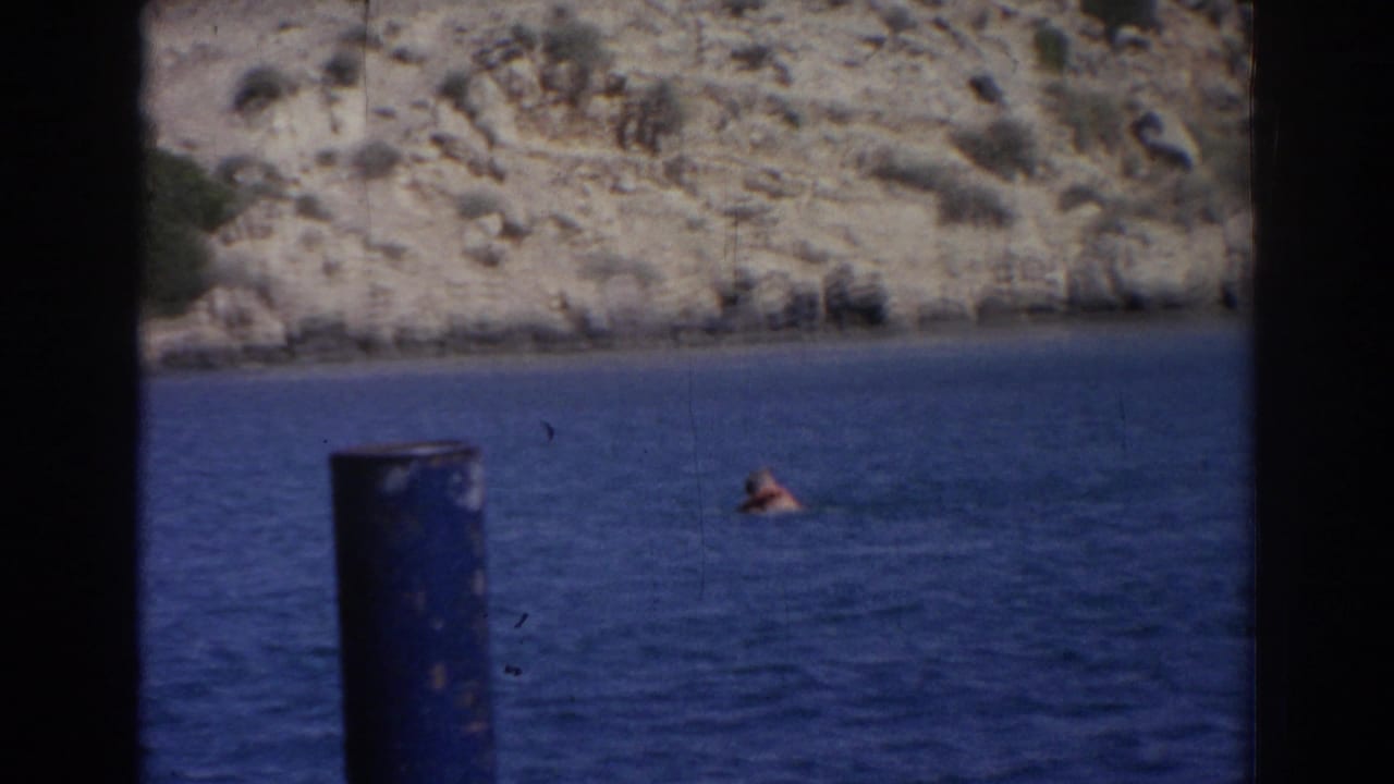 1962 Izmir 8mm frame showing a swimmer offshore, blue water, rocky hillside, and a dark vertical post at the left foreground.