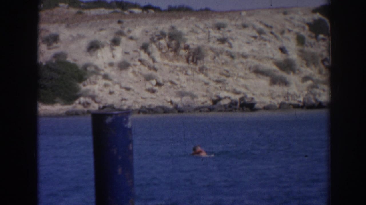 1962 Izmir home-movie frame with a distant swimmer in blue water beneath a sunlit rocky coastal slope.
