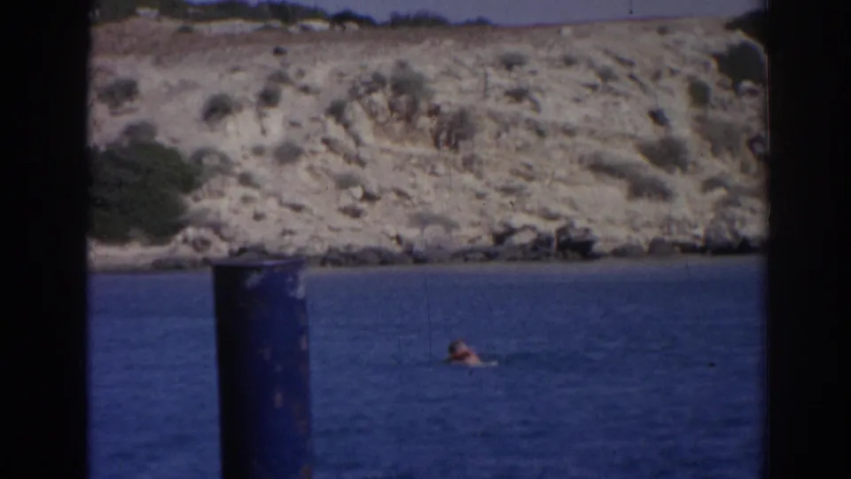 1962 Izmir home-movie frame with a distant swimmer in blue water beneath a sunlit rocky coastal slope.
