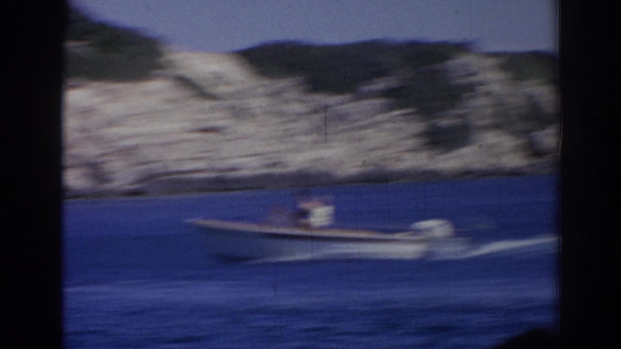 Motion-blurred 1962 Izmir 8mm frame showing a small open motorboat moving across the bay near a rocky coast.