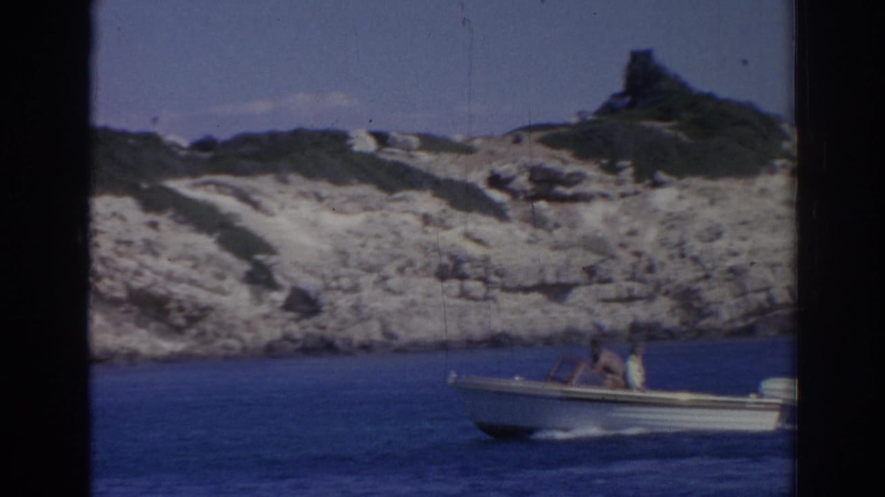1962 Izmir 8mm frame showing a small open motorboat with two occupants passing a rocky shoreline and distant hilltop structure.