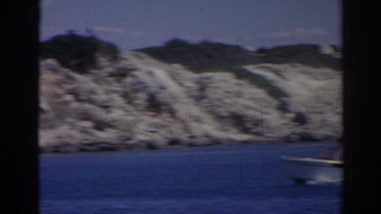 1962 Izmir home-movie frame with a small motorboat on blue water near a sunlit rocky shore.