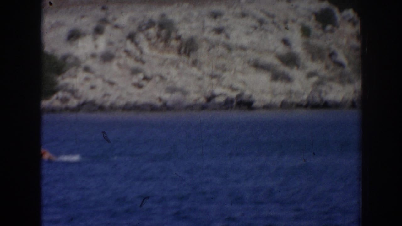 Distant swimmer in red on blue water near the rocky coast in 1962 Izmir footage.
