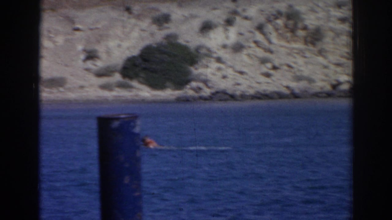 A swimmer in red passes near a dark foreground post, with blue water and rocky Izmir shoreline beyond.