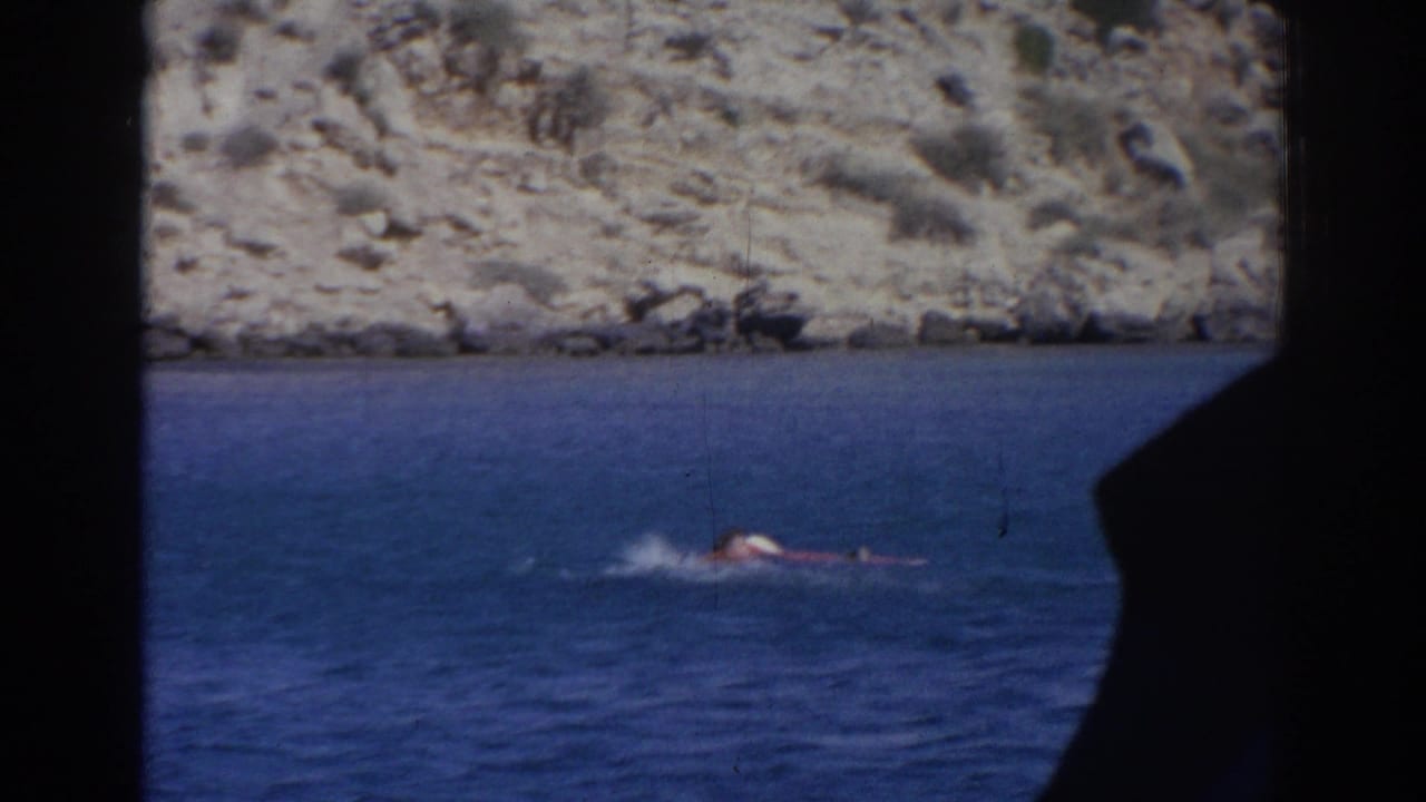 1962 Izmir home-movie still of a swimmer struggling low in blue coastal water beneath a rocky shore.