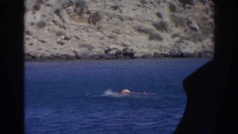 1962 Izmir home-movie still of a swimmer struggling low in blue coastal water beneath a rocky shore.
