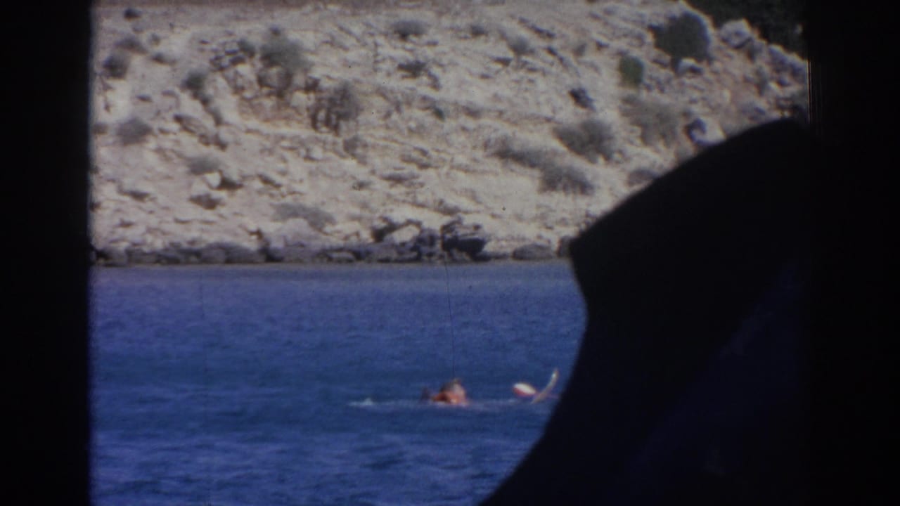1962 Izmir home-movie frame showing a distant swimmer and raised ski on blue water beside a rocky shore, partly framed from inside a boat.