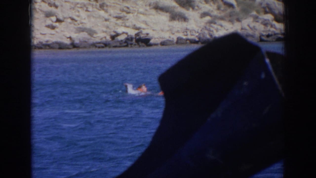 Distant swimmer on blue water in Izmir, Turkey, with rocky shoreline behind and dark boat framing at the right edge.