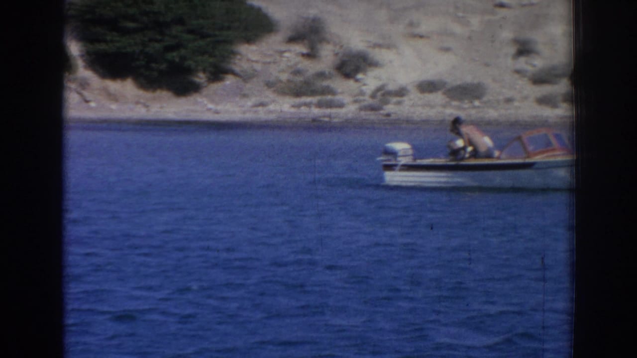 Small motorboat with a person aboard on the water near a rocky Izmir shoreline in a 1962 home-movie frame.