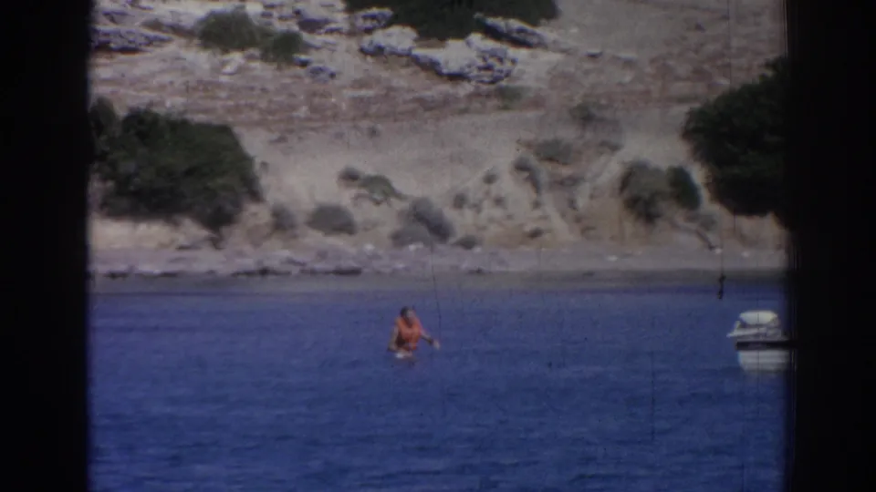 Person in an orange swimsuit wading or swimming near a rocky Izmir shoreline, with a small boat partly visible at frame edge.