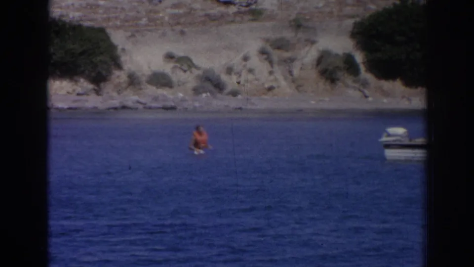 Distant swimmer in blue water below the rocky Izmir shoreline, with a small boat cropped near the right edge of the 8mm frame.