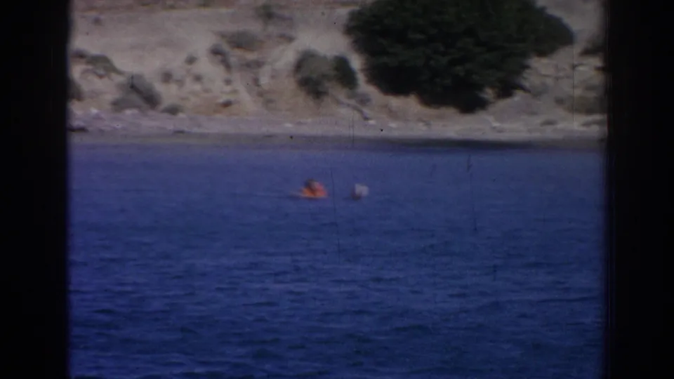 A swimmer appears farther from camera in blue water near the rocky Izmir shoreline, with a small pale object or splash nearby.