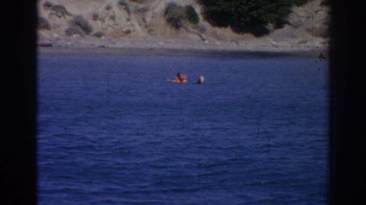 Distant person in orange life vest floating in blue water near a rocky shore.