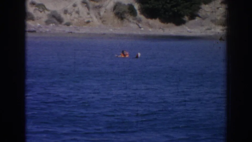 1962 Izmir home-movie frame showing a distant person in an orange life vest in deep blue water near a dry, rocky shoreline.