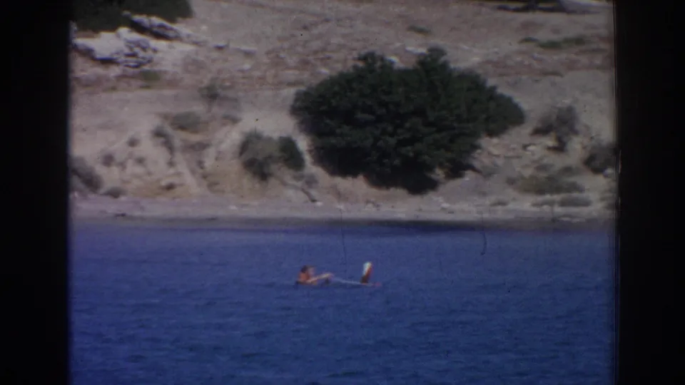 1962 Izmir 8mm frame showing a distant person near a white board or float, with scrub and rocky shoreline above the water.