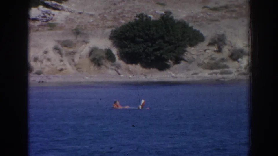 1962 Izmir coastal home-movie frame with a person in an orange life vest beside a white float or board under bright sun.