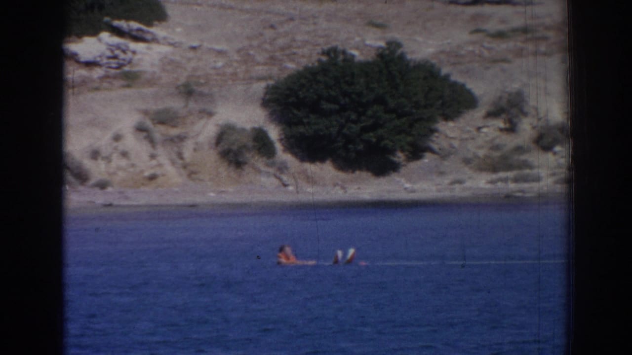 Distant person floating in blue water with water skis near a dry rocky shoreline.