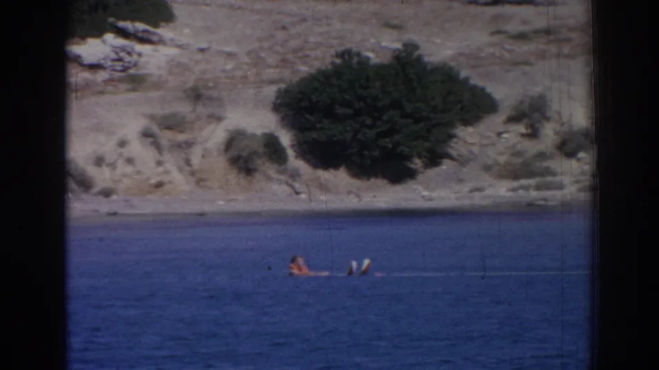1962 Izmir home-movie frame showing a person waiting in blue water beside water skis, with a rocky shrub-lined shore behind.