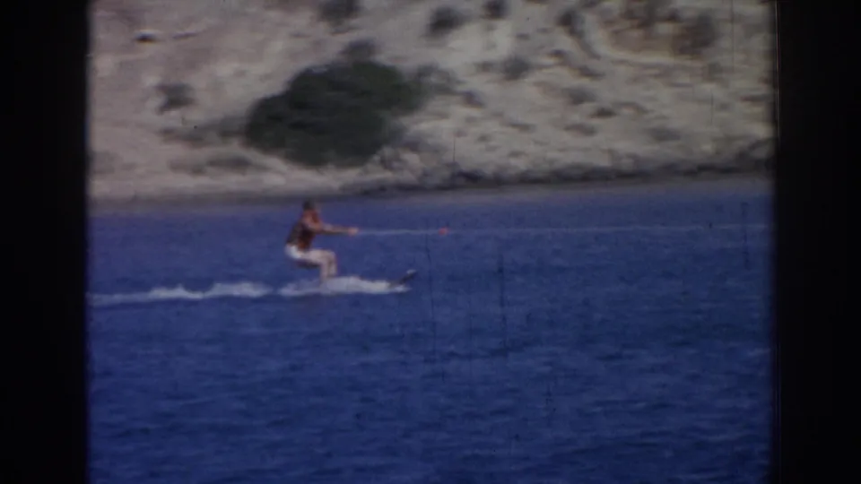 1962 Izmir frame showing a skier upright on water skis, pulled by a tow rope across blue water near a dry shoreline.