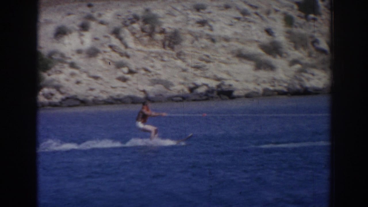 Male water skier crosses blue water with a rocky shoreline behind him.