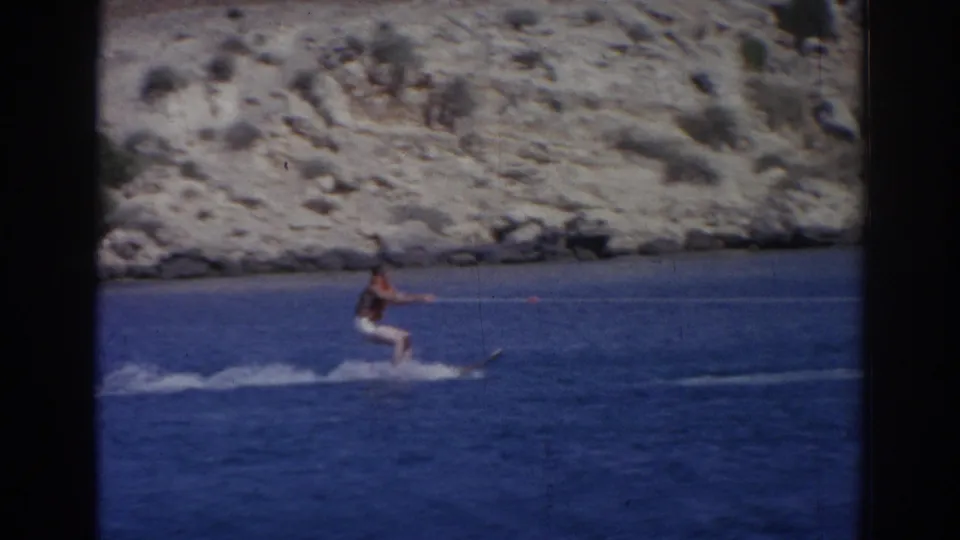 1962 Izmir home-movie frame showing a male water skier being towed across deep blue water near a rocky coast.