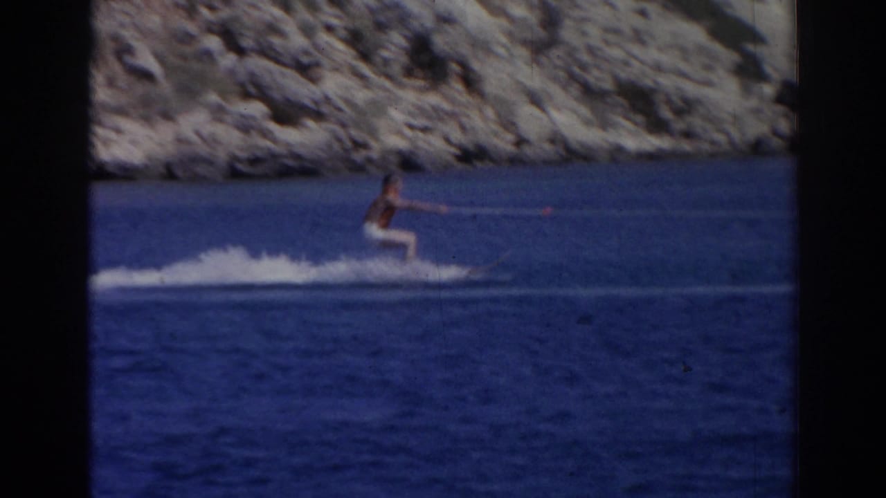 Male water skier in 1962 Izmir footage glides across blue water while holding a tow rope near a rugged shoreline.