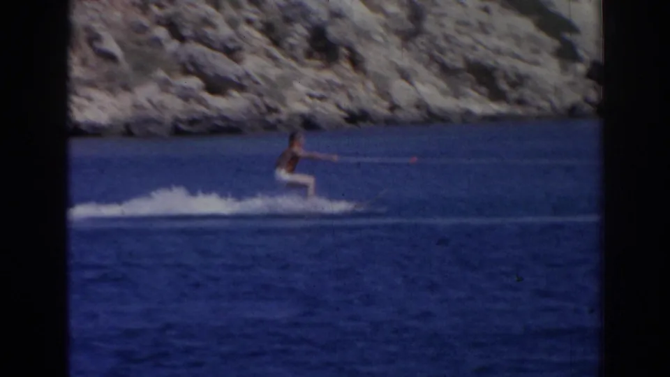 Male water skier in 1962 Izmir footage glides across blue water while holding a tow rope near a rugged shoreline.